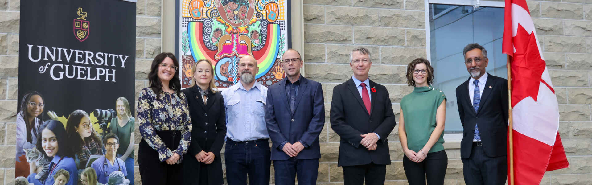 a group of men and women pose for a photo between a university of guelph sign and a canadian flag