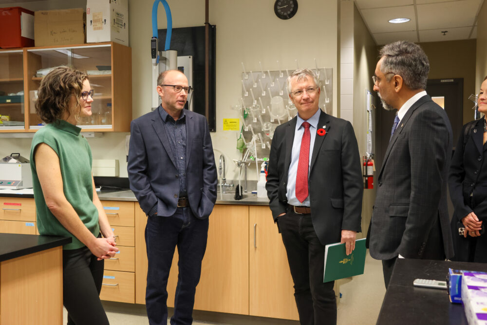 a woman in a green shirt and three men in suits stand in conversation in a lab setting