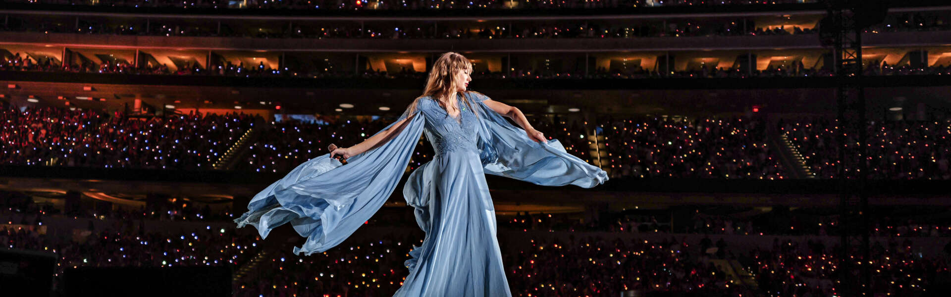 A woman in a blue dress with flowing sleeves stands on stage in front of a crowd of thousands backlit with stadium lighting.