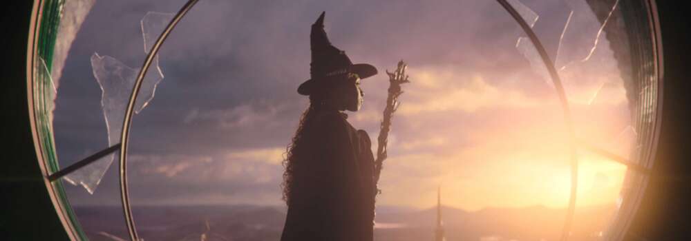 a woman in a black cape and hat stands in front of a circular window, she's holding a broom