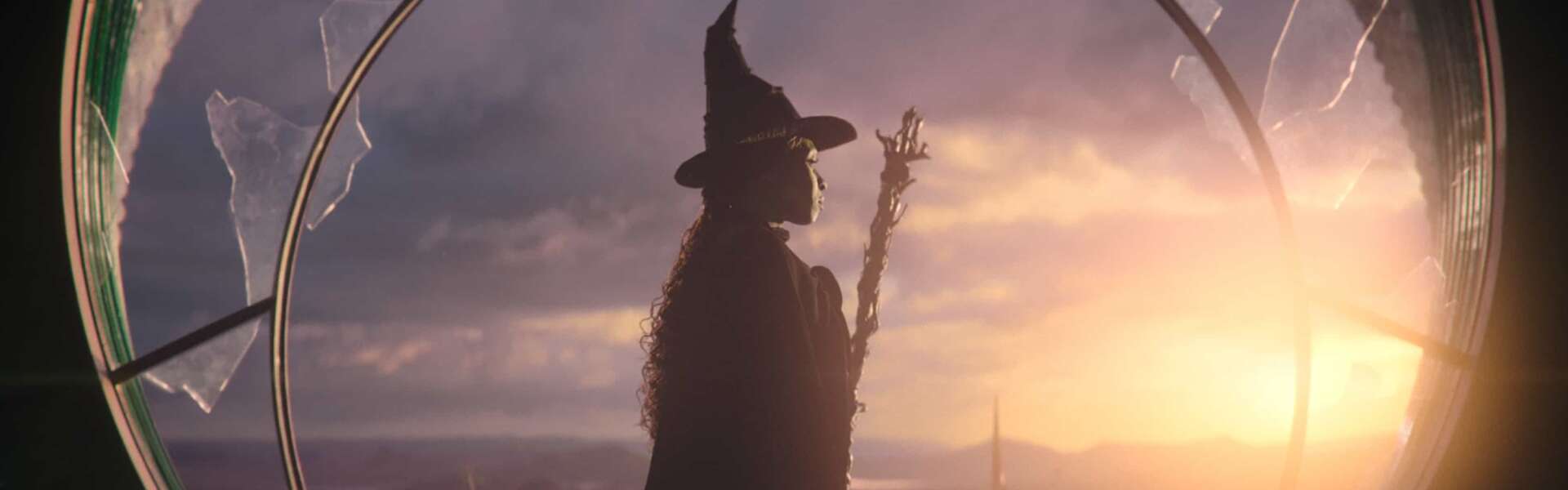 a woman in a black cape and hat stands in front of a circular window, she's holding a broom