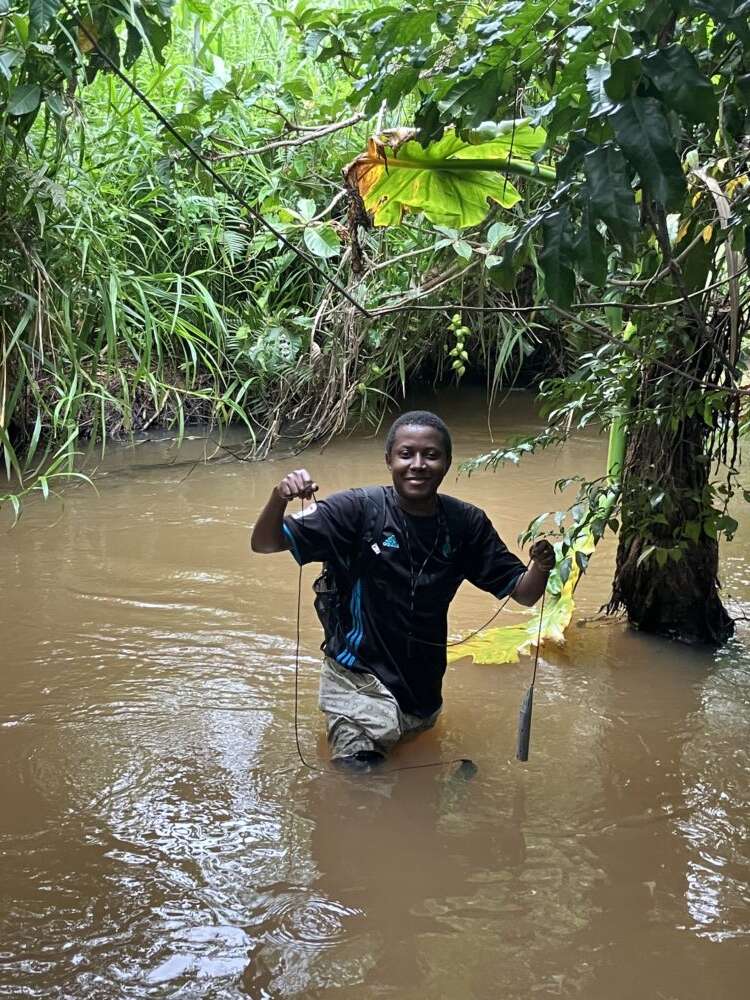 Amilcar Bisekere standing knee-deep in a muddy river, surrounded by lush greenery, wearing a black shirt and light shorts, holding sampling equipment while smiling at the camera
