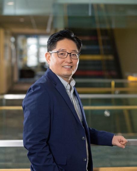 a man in a blue suit jacket with black hair and glasses poses for a headshot in front of a staircase