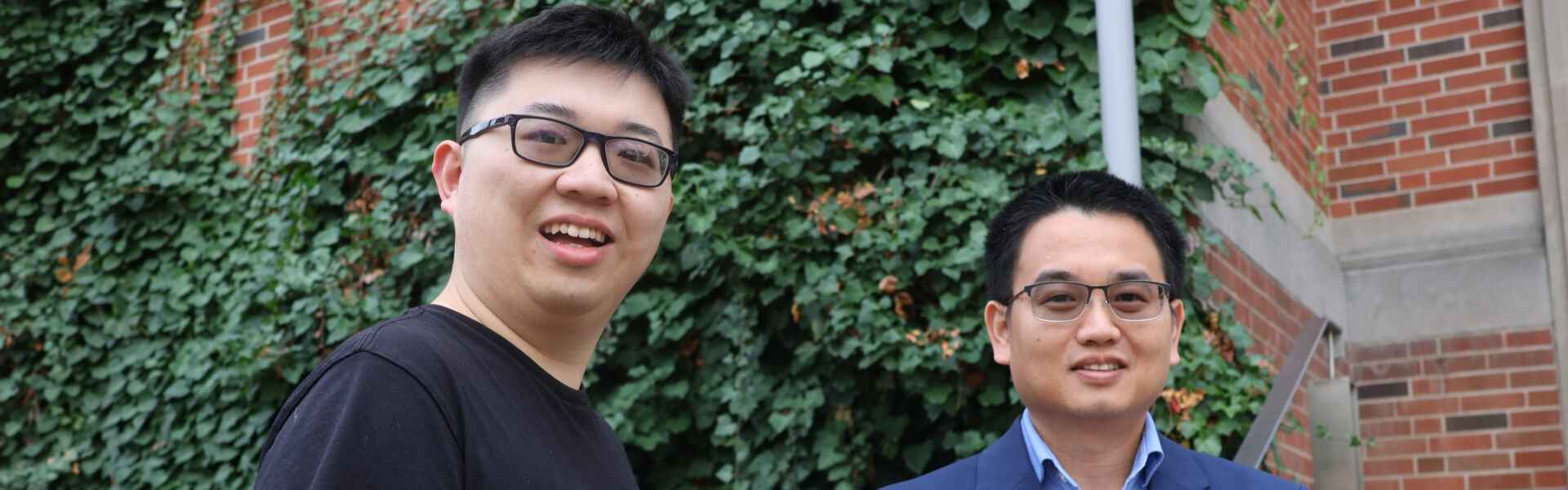 Two people with glasses, on wearing a black t-shirt and one wearing a blue button-up under a blue blazer stand in front of greenery and a red brick wall.