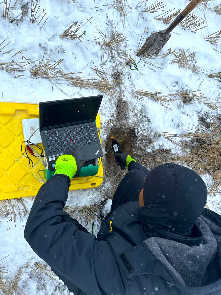 Overhead shot of a student applying a device to the ground in the middle of winter, a laptop connects the device as they analyze the data