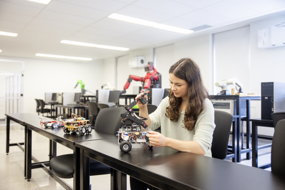 Student on a desk playing with a robotic prototype, looking intensely at a wheel and arm component