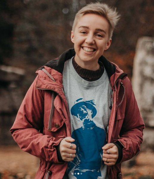 a person with short blonde hair wearing a grey t-shirt and a rust jacket poses for a headshot