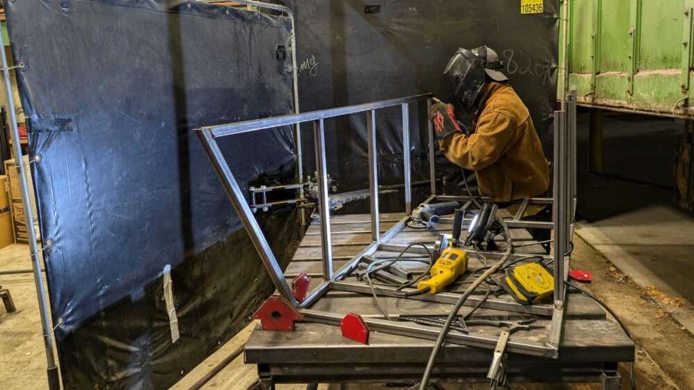 a student in a brown jacket wearing a black helmet works on the frame of a toboggan