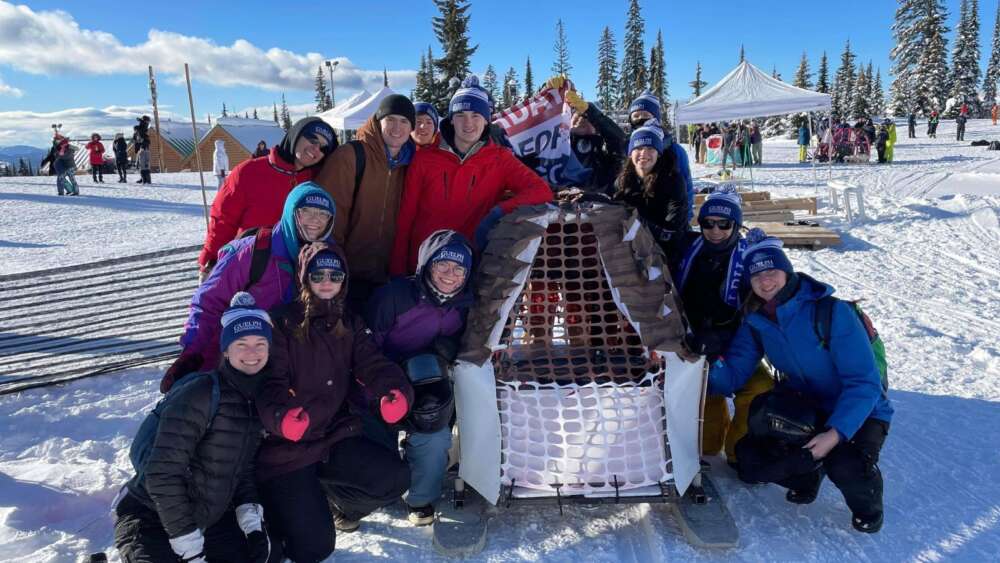 students in winter jackets pose in the snow for a group photo with a toboggan