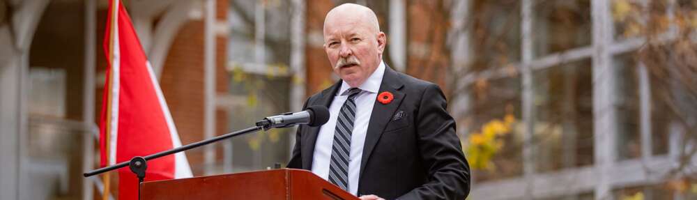 a man in a suit wearing a poppy speaks at a podium in front of a Canadian flag