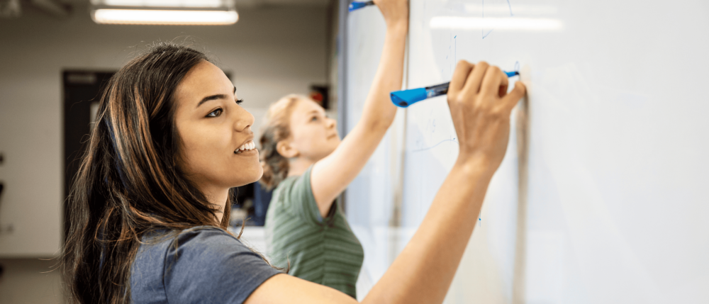 Two students draw on a white board