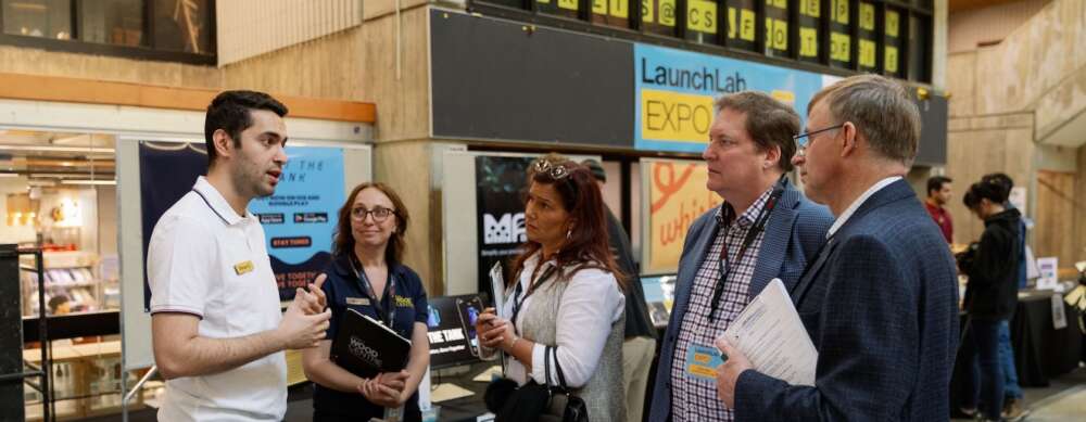 A group of five people stand around a table showcasing a product while one of them talks to the others about their work in the UC during the LaunchLab Expo.