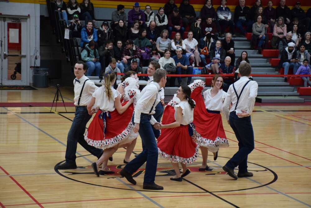 dancers in white shirts, black pants and red shirts dance on a gym floor