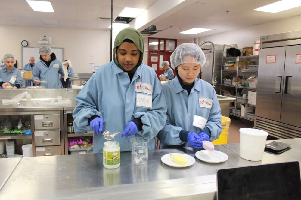 students in blue lab coats and blue gloves scoop ingredients using spoons at a lab bench
