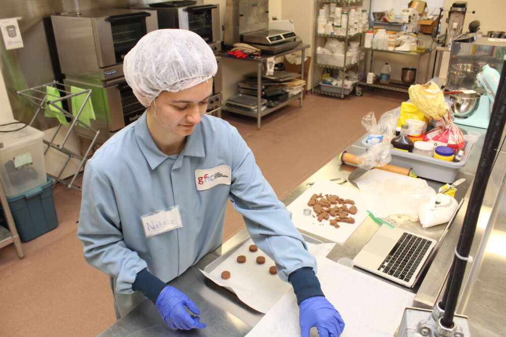 a student in a blue lab coat, blue gloves and a white hairnet places a food item on a piece of white paper