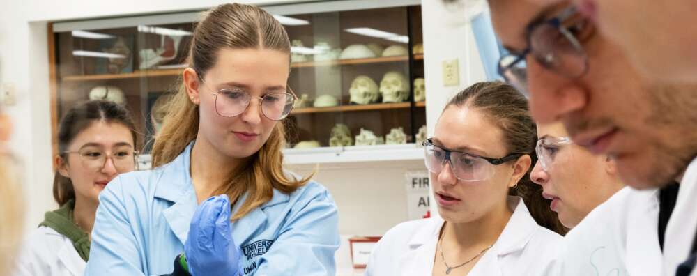 A group of students in white lab coats, goggles and blue latex gloves stand listening to an instructor in a blue lab coat and goggles.