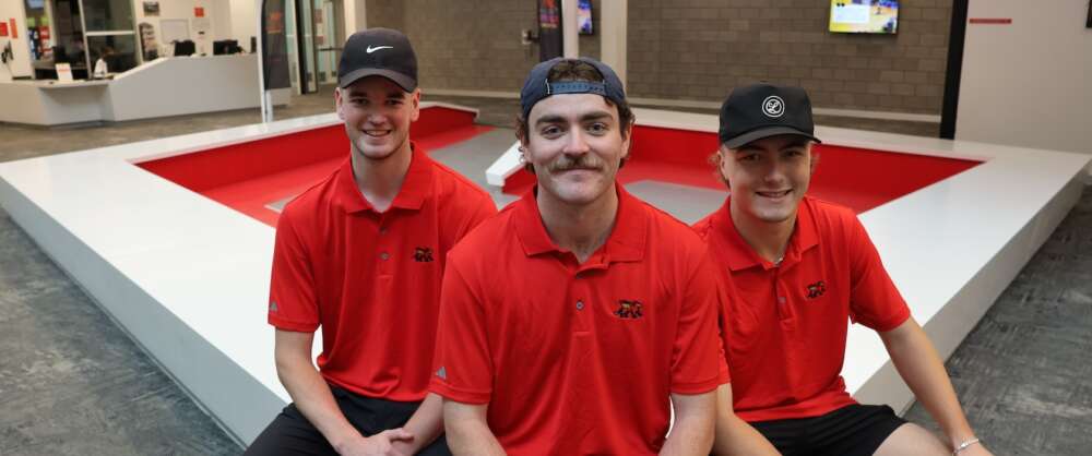 Three students in black hats and red U of G golf shirts sit forming a tripod and smiling into the camera in the lobby of the Athletic Centre