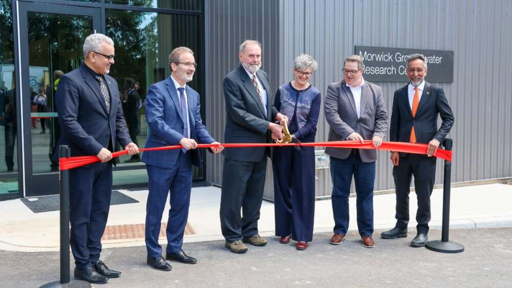 Six people hold a red ribbon in front of a steel covered building