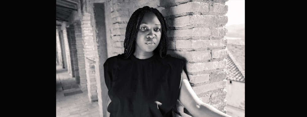 A person with long, black braids wearing a black sleeveless t-shirt stands against a brick wall with one arm extended on a ledge.