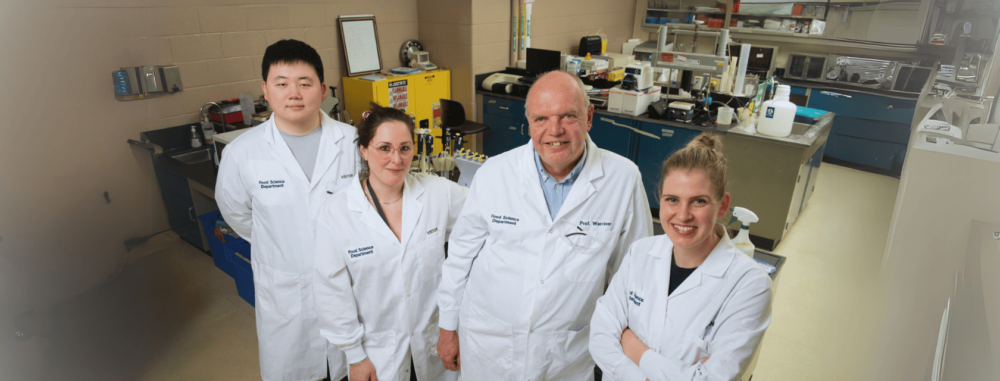 Four food science researchers, including Prof. Warriner, pose together in a lab. They are all wearing white lab coats labeled “Food Science Department.” Lab benches, scientific instruments, and storage cabinets are visible in the background.