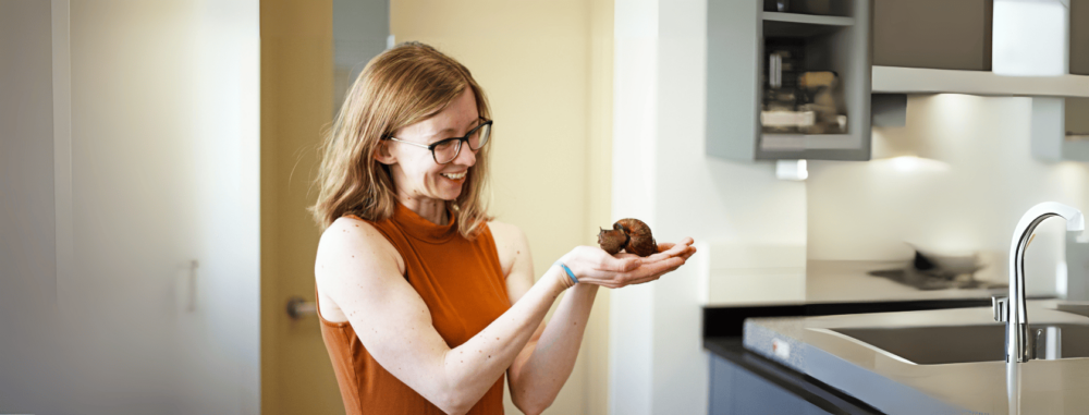 Dr. Sarah Schorno, in glasses and a sleeveless orange shirt, holds a large brown snail in both hands while standing in a lab. She looks down at the snail with fascination and delight