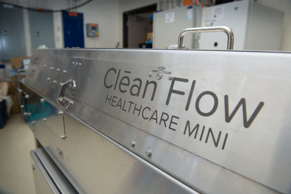 Close-up of the side of a large stainless steel machine labeled “Clean Flow Healthcare Mini.” The machine is located in a laboratory with various equipment and storage cabinets in the background
