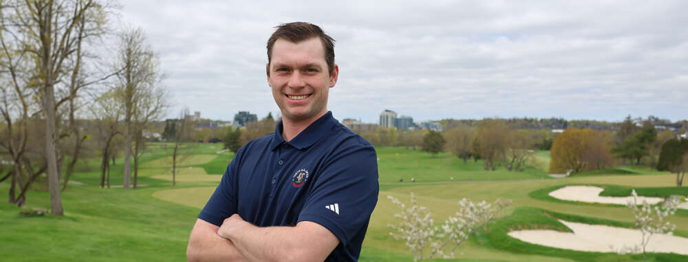 A man with brown hair wearing a navy golf shirt stands smiling with his arms crossed in front of him with a golf course and buildings in the background.