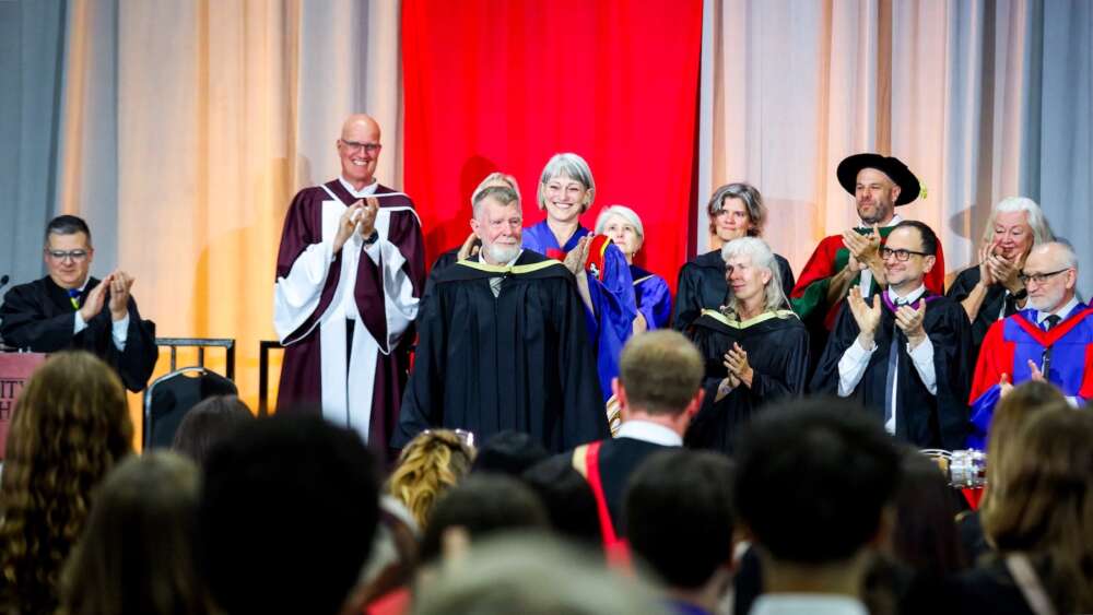 A man with a beard and gray hair wearing a black graduation gown stands on a convocation stage to receive his degree with a group of University leadership standing behind him and an audience in front of them.