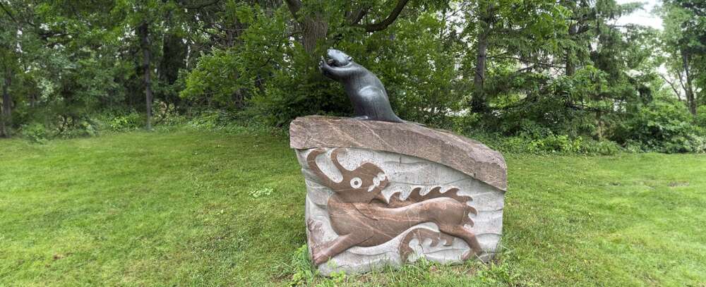 A sculpture depicting a bronze beaver on an engraved granite block, amid a grassy lawn.