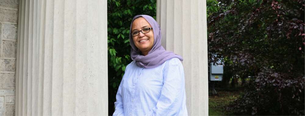 Maysa Niazy, in a lavender headscarf and light blue striped shirt smiles while standing between two large stone columns. Behind her, green foliage and trees fill the background, creating a peaceful and welcoming atmosphere.