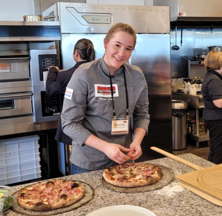 a person stands in a restaurant kitchen adding toppings to a pizza