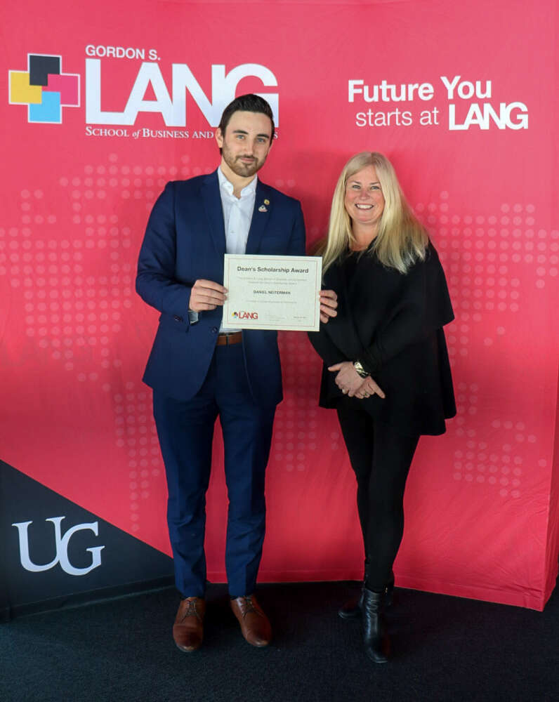 A young man in a navy suit holds a Dean’s Scholarship Award certificate while standing next to a smiling woman in black. They pose in front of a red University of Guelph Lang School of Business and Economics backdrop with the words “Future You starts at LANG.”