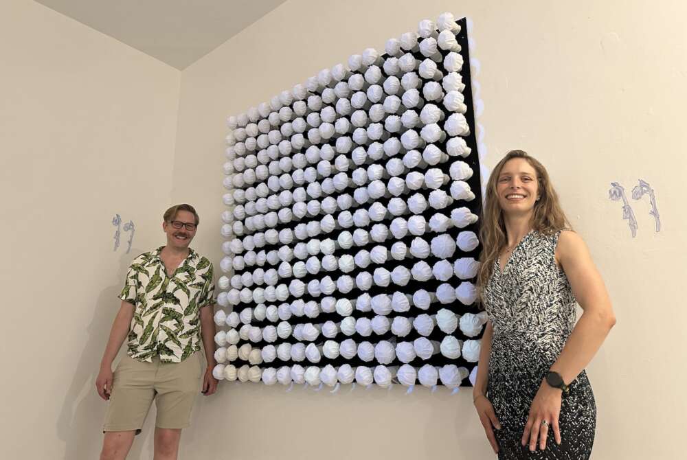 A person in a leaf patterned button-up shirt snd beige shorts with a mustache and glasses stands beside a piece of black and white art mounted on a wall while a second person in a black and white patterned dress with long brown hair stands on the other side.