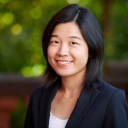 a person with black in a white shirt and navy blazer poses for a headshot in front of a green background