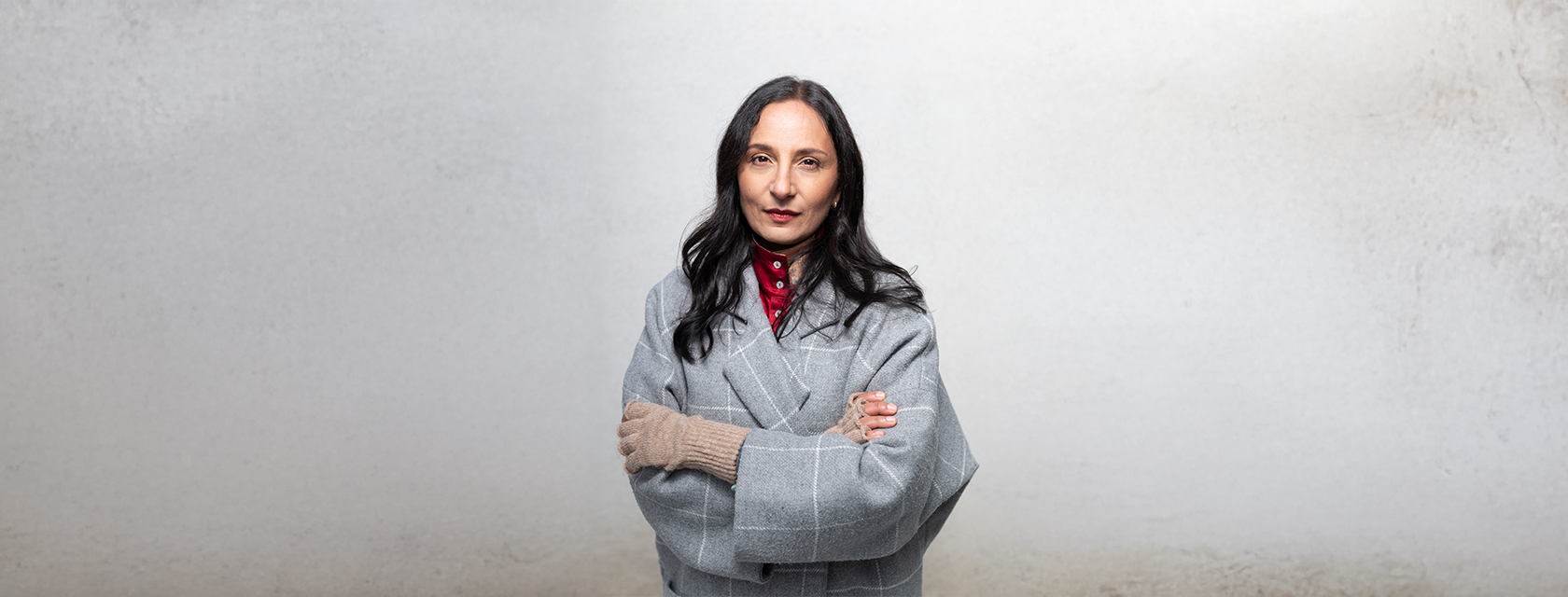 Madhur Anand, dressed in a gray coat, crosses her arms powerfully against a gray background
