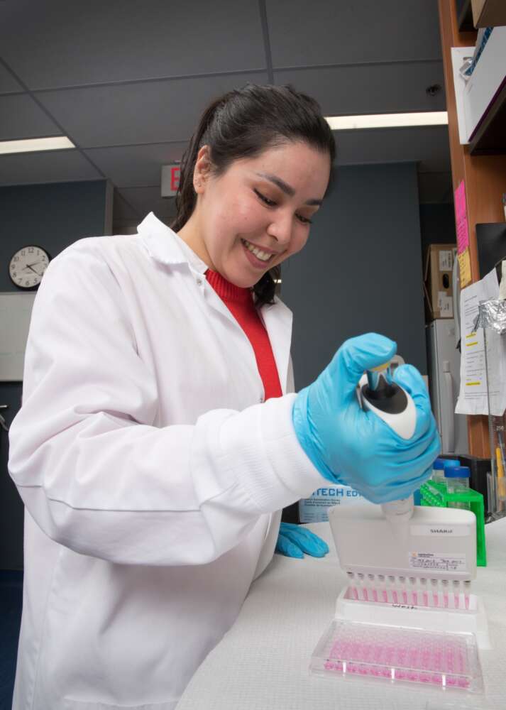 A researcher in a white lab coat and blue gloves uses a multi-channel pipette to transfer liquid into a 96-well plate containing pink fluid. She is smiling and focused on her work inside a laboratory, surrounded by various lab supplies and equipment