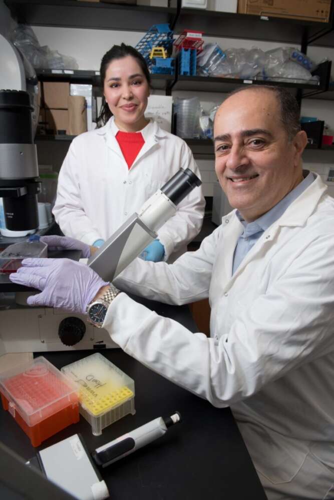 Two researchers in lab coats, a woman and a man, pose in a laboratory. The man, seated at a microscope, smiles toward the camera while wearing gloves and a wristwatch. The woman stands behind him, also smiling, with gloved hands and a red shirt under her lab coat. Lab equipment, including pipette tip boxes and supplies, are visible on the bench and shelves.