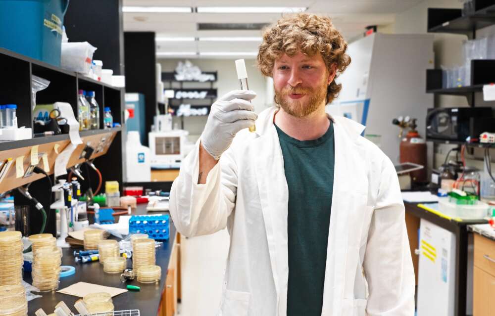 A person with curly hair and a beard wearing a green t-shirt under a white lab coat holds a tube with a gloved hand and peers into its contents.