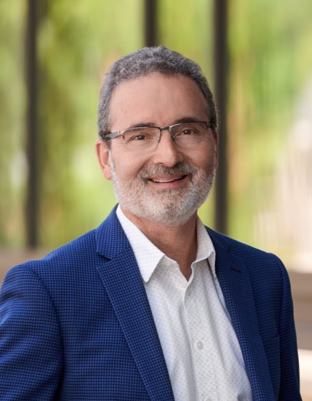 Dr. Rene Van Acker, dressed in a navy blue suit and white dress shirt, stand against a fence area on a clear sunny day