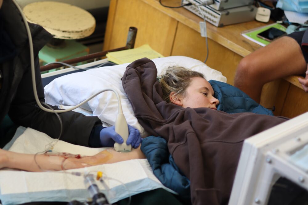 A person lies on a bed covered in a sleeping bag while researchers conduct tests on them using ultrasound equipment.