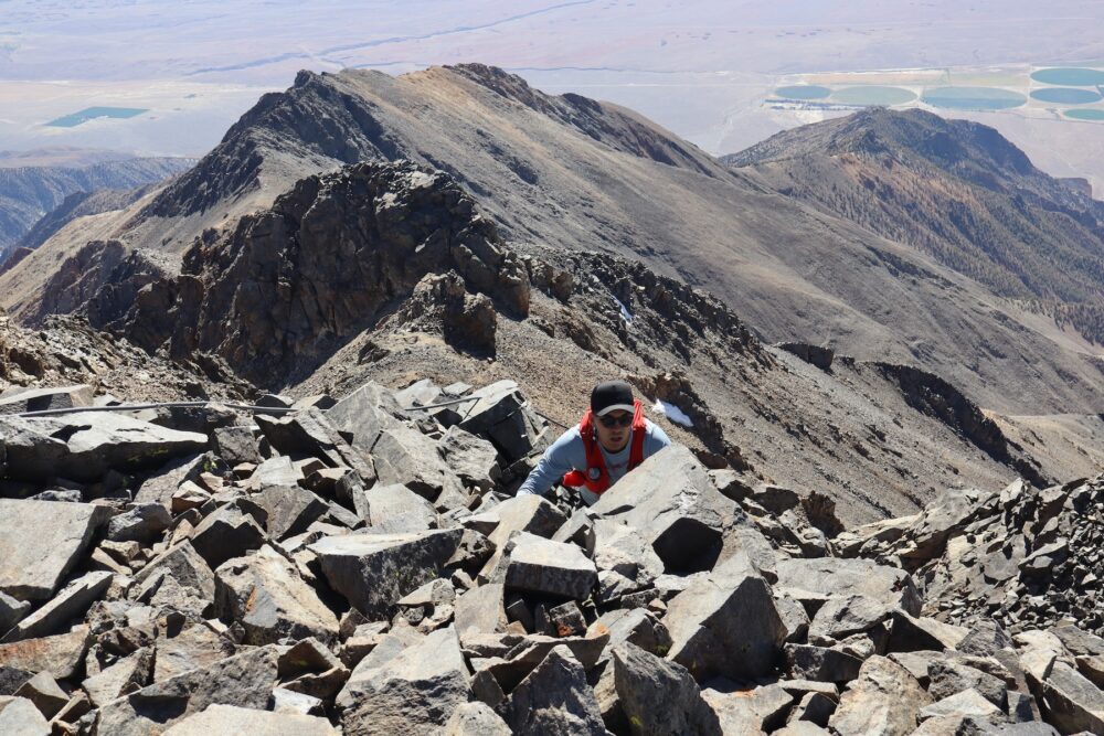 A person wearing a black hat, blue t-shirt and red backpack climbs over rocks, toward the camera, at the peak of a mountain.