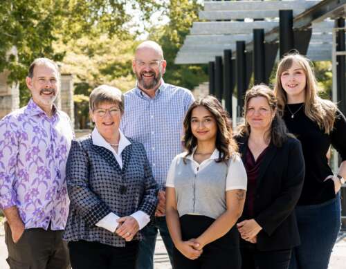A group of six people stand staggered wearing a mix of colours and smiling for the camera in front of a glass building and some greenery.