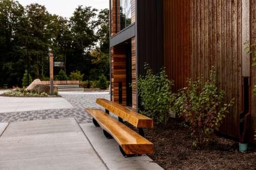 A wooden bench is pictured outside a newly restored wooden building with some shrubs and a concrete sidewalk leading inside.