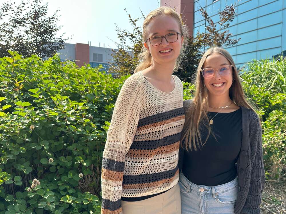 a person in a brown striped sweater and a person in a black t shirt and cardigan pose for a photo in front of a green background
