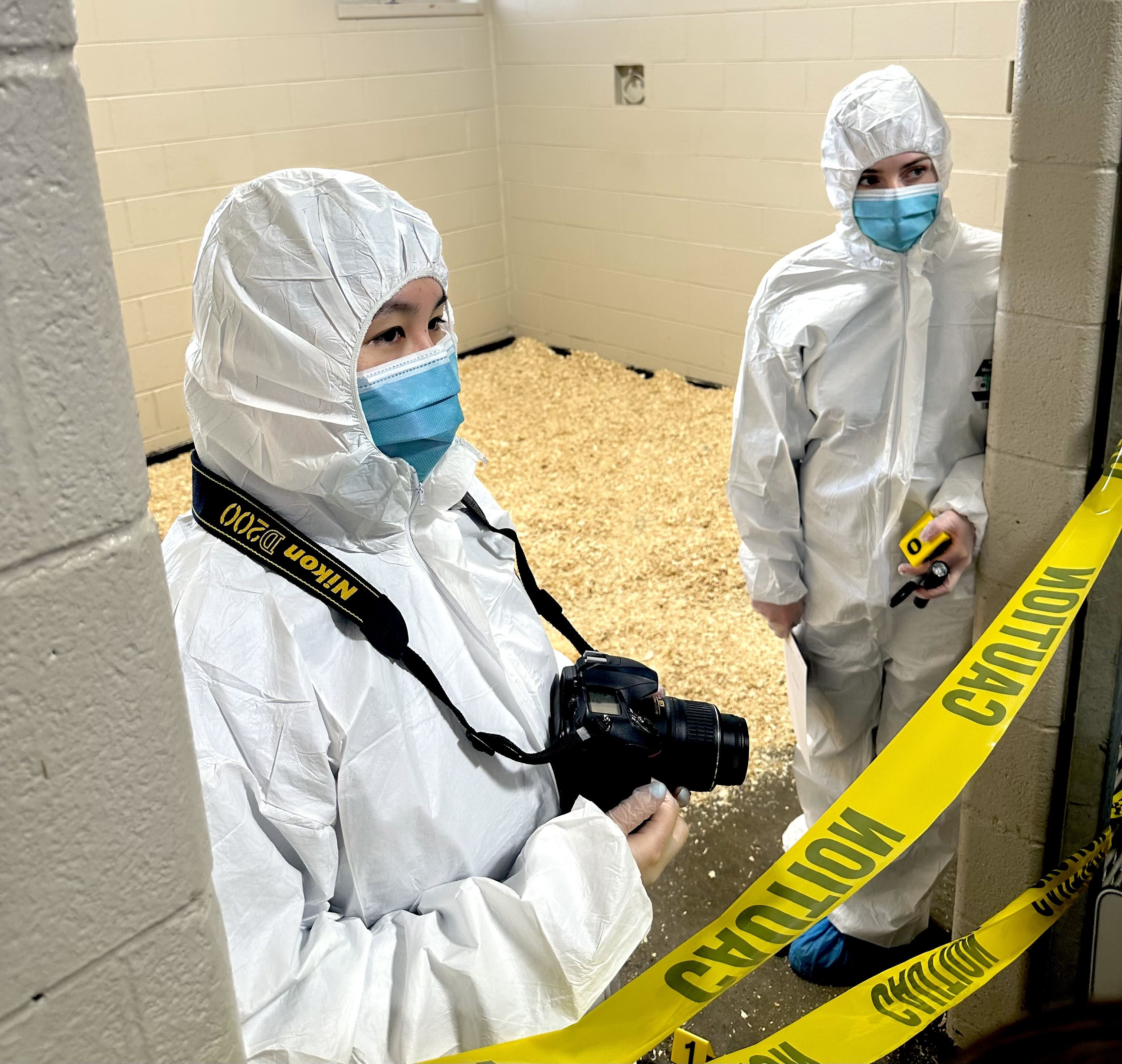 Two individuals wearing full white protective suits, blue surgical masks, and shoe covers stand behind yellow caution tape in a sterile room with wood shavings on the floor. One person holds a camera and the other holds measuring tools, suggesting a forensic investigation scene.
