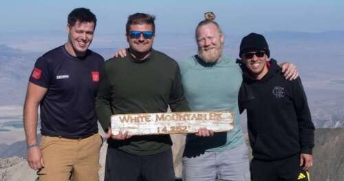 Four researchers stand together at the summit of White Mountain holding a white sign with text that reads the elevation of the mountain.