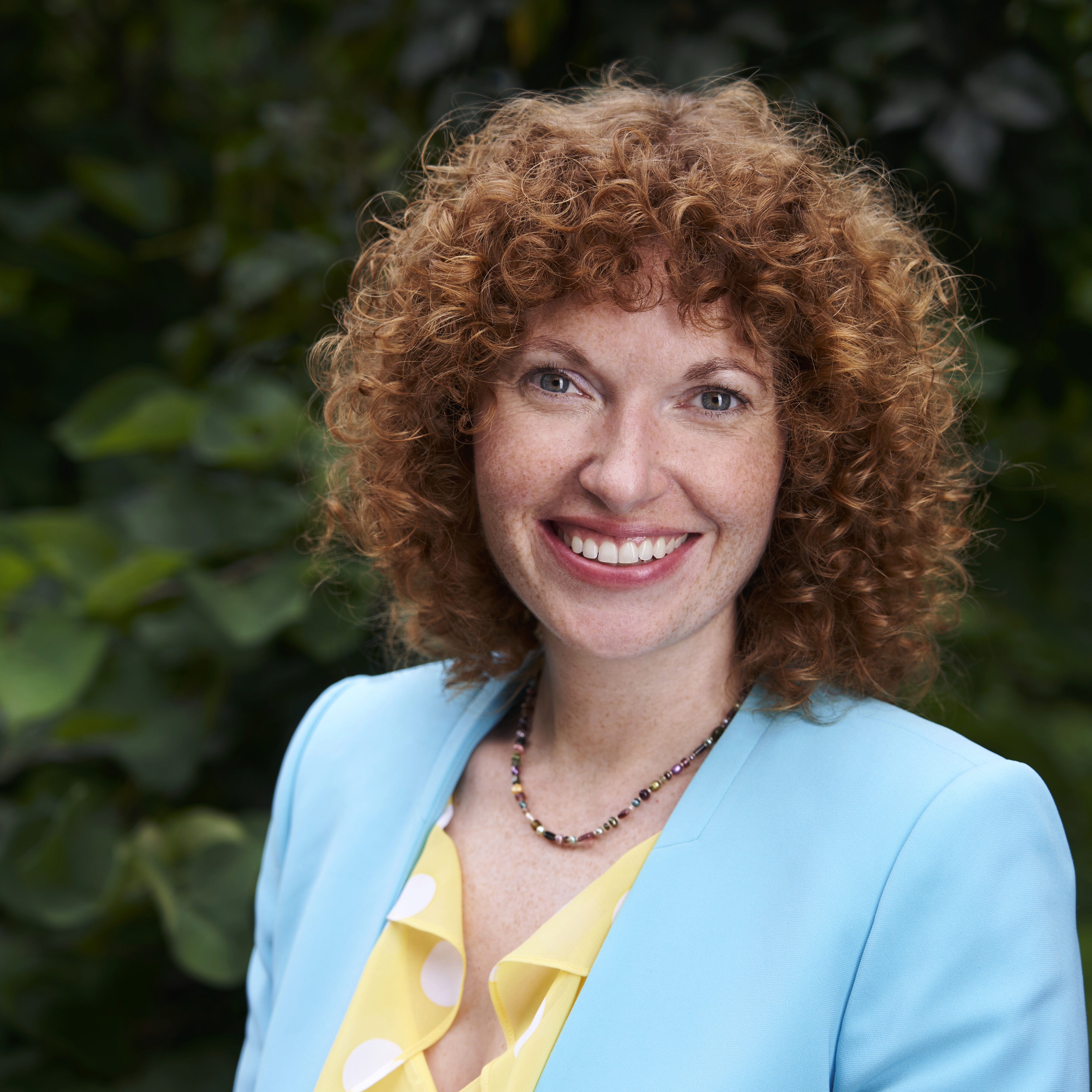 a person with curly red hair in a blue blazer and yellow shirt poses for a headshot