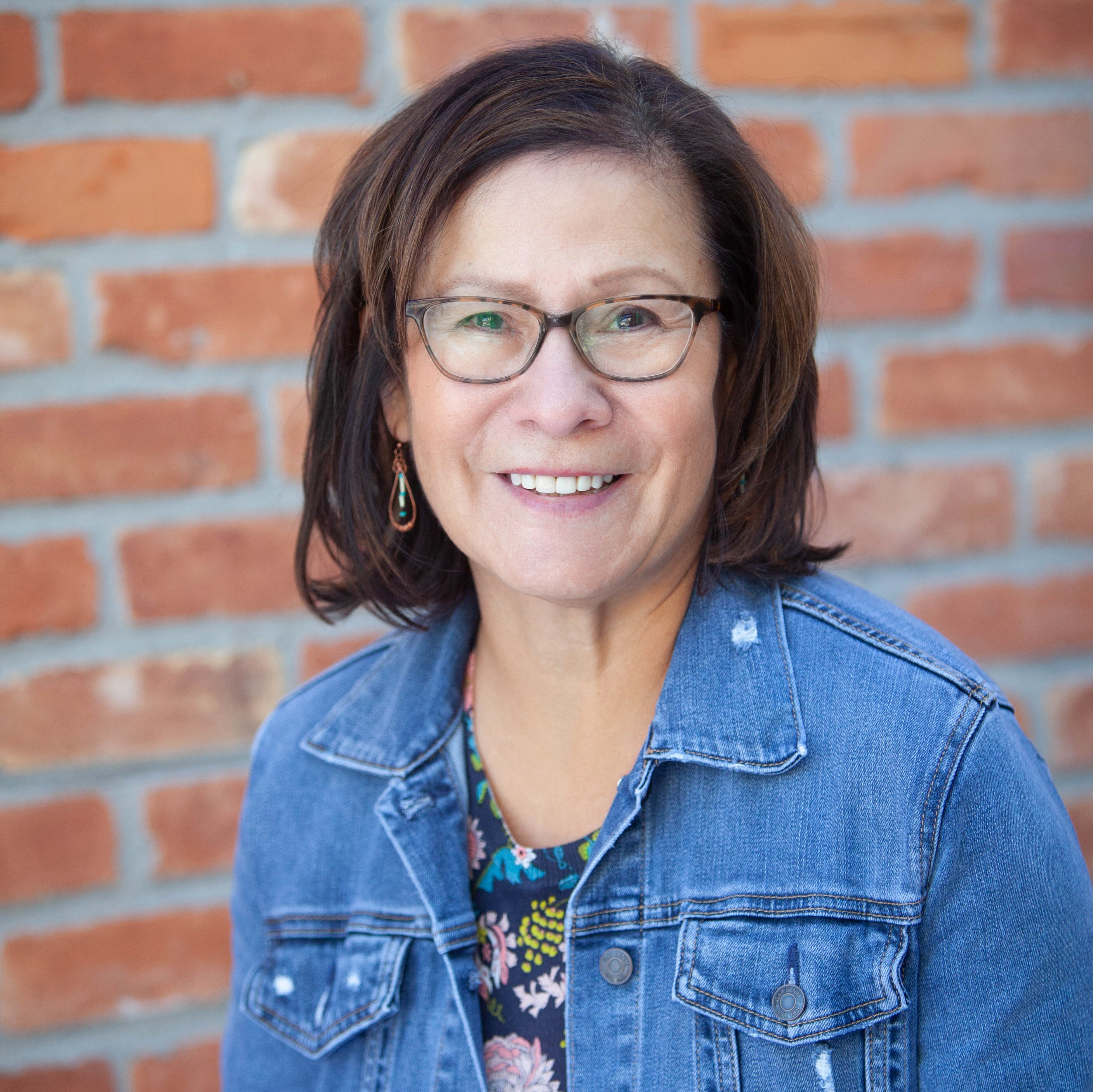 a person with long dark hair and glasses wearing a blue denim jacket and a floral shirt poses for a headshot in front of a red brick wall