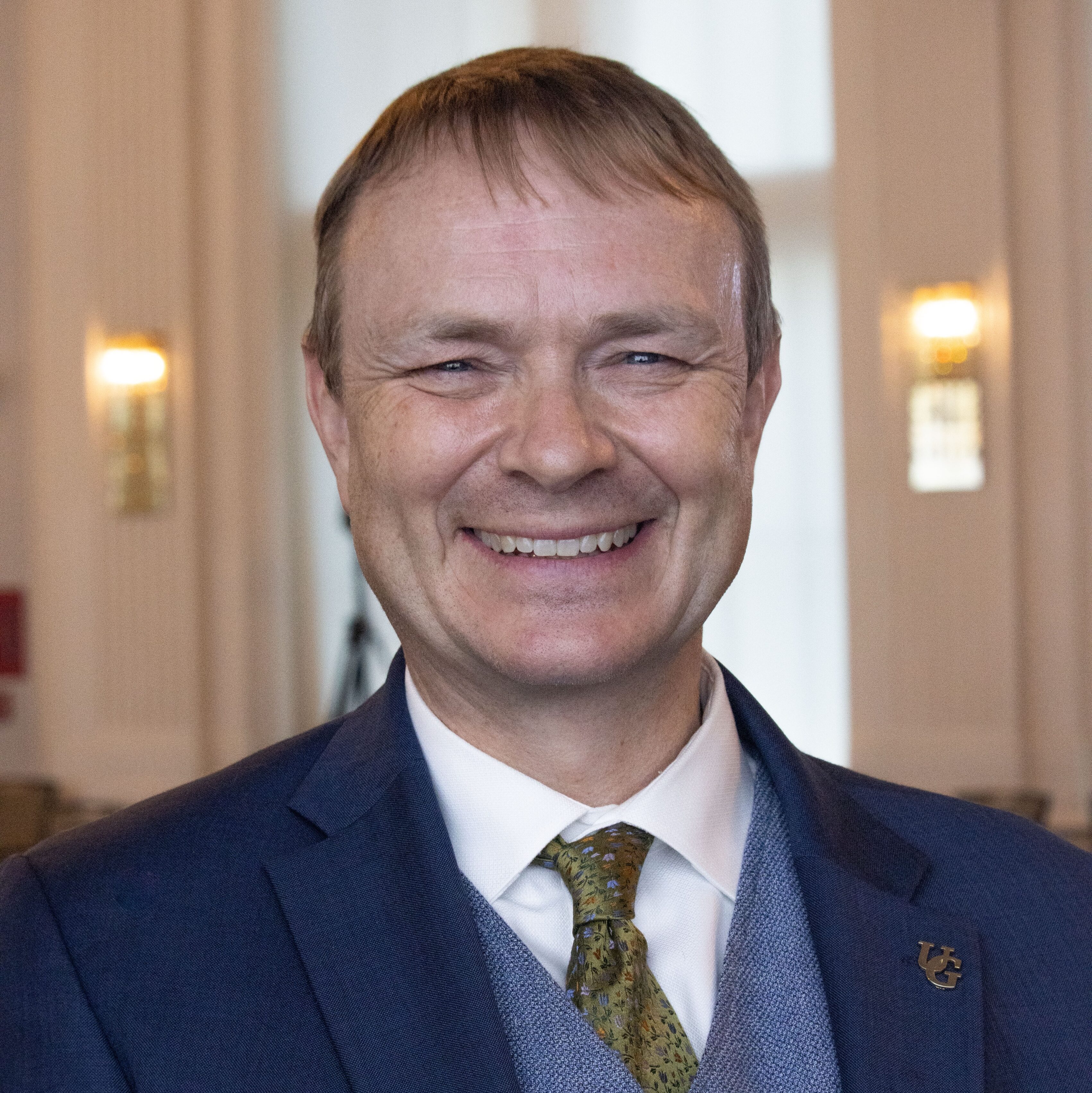 a person with short hair wearing a navy blue jacket and blue vest poses for a headshot