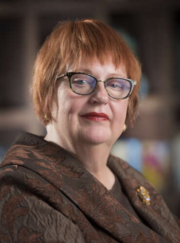 a person with short red hair and glasses wearing a brown jacket poses for a headshot